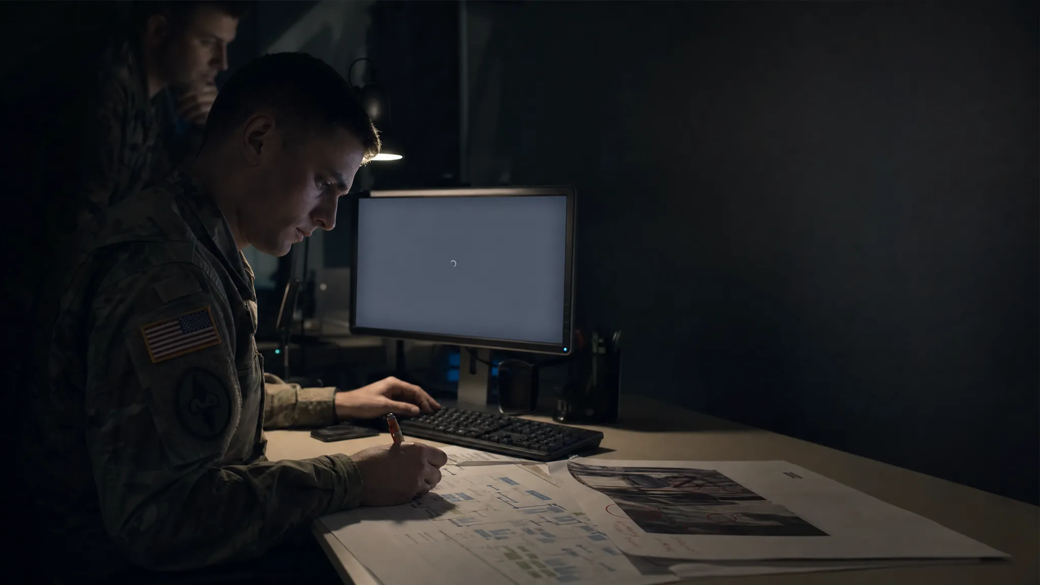 Landscape late-evening office scene with a forward deployed engineer seated left-of-center, studying a printed customer workflow map and a marked-up output sample spread across the desk. A monitor faces the camera and shows a mostly blank interface with a waiting cursor and no other content. One hand rests near the keyboard while the other holds a pen over a correction on the page. A second person is faintly visible in the background, standing and looking at the same desk from over the engineer’s shoulder. Practical desk lamp and monitor glow only, deep clean shadows, subdued cool-toned accent, quiet and unresolved mood. Keep the right side of the frame open for text overlay.